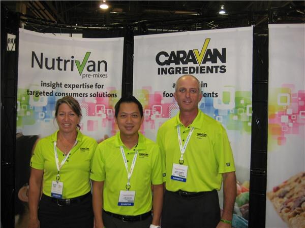 Two men and a woman in lime green polo shirts and exhibitor badges stand in front of Nutrivan pre-mixes and Caravan Ingredients banners at a trade show booth.