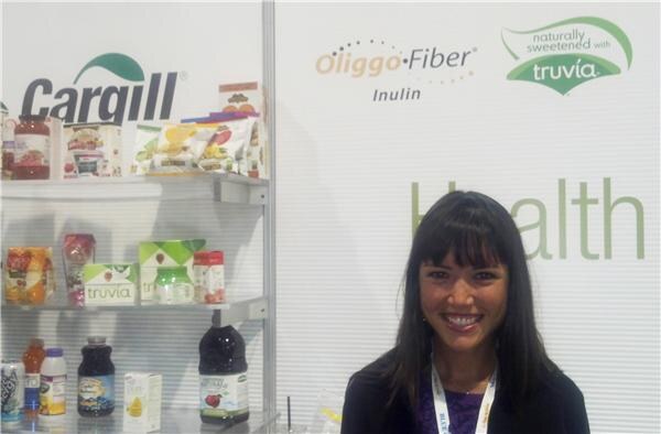 A smiling woman with dark hair and bangs stands in front of Cargill shelves of Truvia products and signs for Oliggo-Fiber Inulin and Truvia.