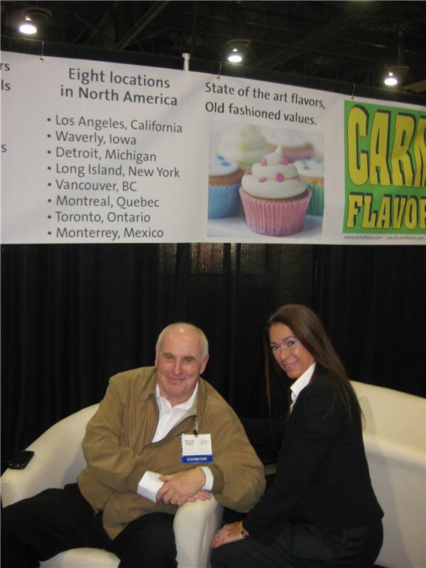 A man in a tan jacket and a woman in black sit on a white sofa under a Carmi Flavors banner listing eight North American locations and showing cupcakes.