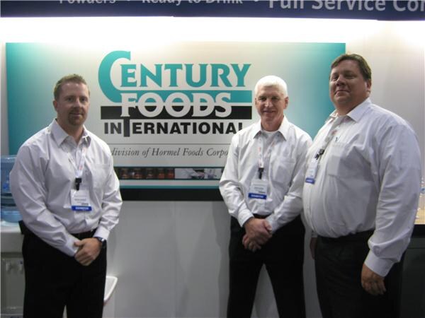 Three men in white dress shirts and exhibitor badges stand in front of a Century Foods International sign, a division of Hormel Foods Corporation.
