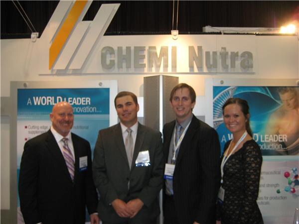 Three men in dark suits and a woman in a black lace dress stand together beneath a CHEMI Nutra sign with blue banners reading A World Leader.