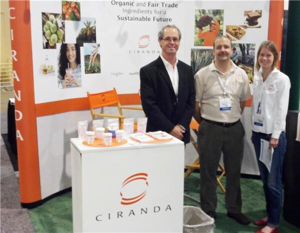 Two men and a woman stand at a white Ciranda display counter with sample jars, in front of banners about organic and fair trade ingredients.