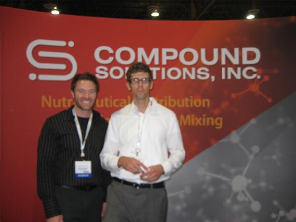 Two men stand in front of a red Compound Solutions, Inc. backdrop; one wears a black striped shirt and an exhibitor badge, the other a white shirt and glasses. The photo is blurry.