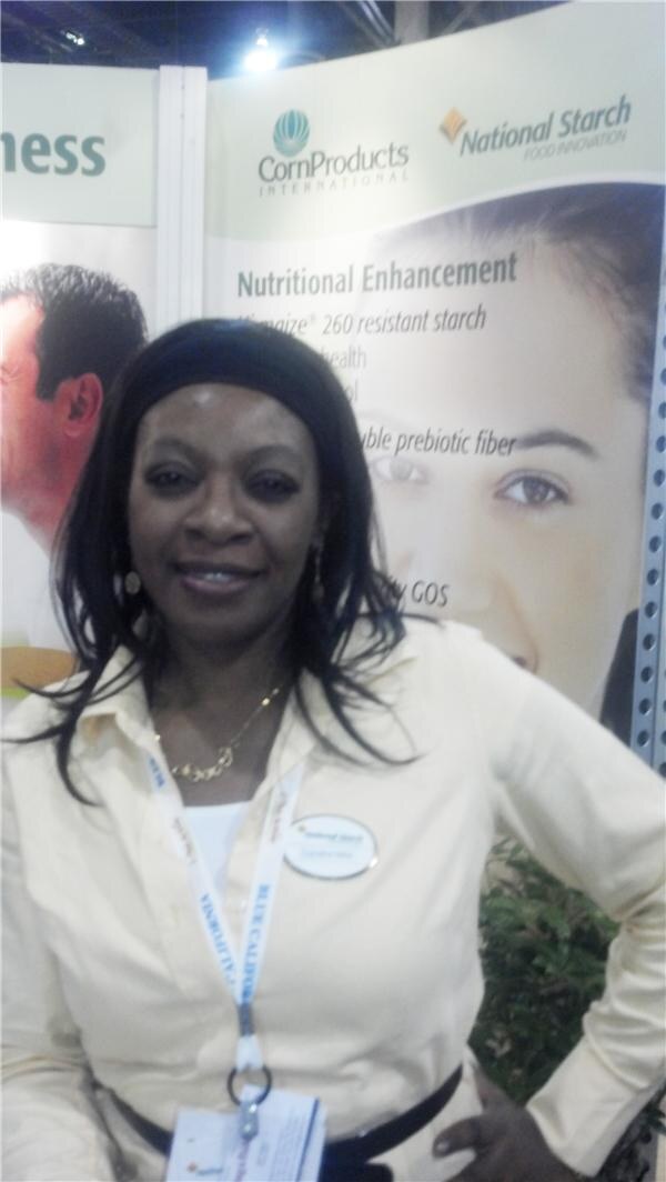 A woman in a pale yellow shirt with a National Starch name tag stands with her hand on her hip in front of a CornProducts International and National Starch display.