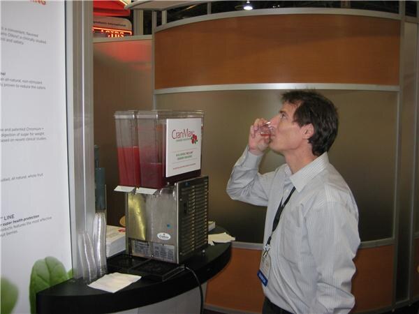 A man with a lanyard drinks from a small cup beside a two-tank dispenser of red juice labeled CranMax, set on a black counter in a booth.