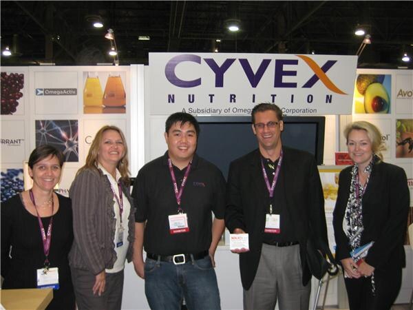 Two men and three women wearing lanyards stand in front of a Cyvex Nutrition sign; one man holds a small product box.