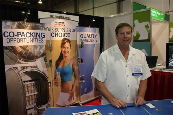 A man in a white short-sleeved shirt and exhibitor badge stands at the Dairy Farmers of America booth beside banners reading Co-Packing Opportunities, Your Supplier of Choice and Quality Ingredients.