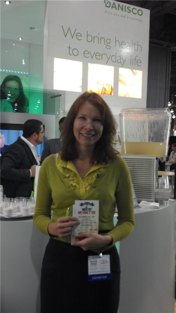 A smiling woman with an exhibitor badge holds a card and pen in front of a Danisco display reading 'We bring health to everyday life', with a beverage dispenser and small cups nearby.