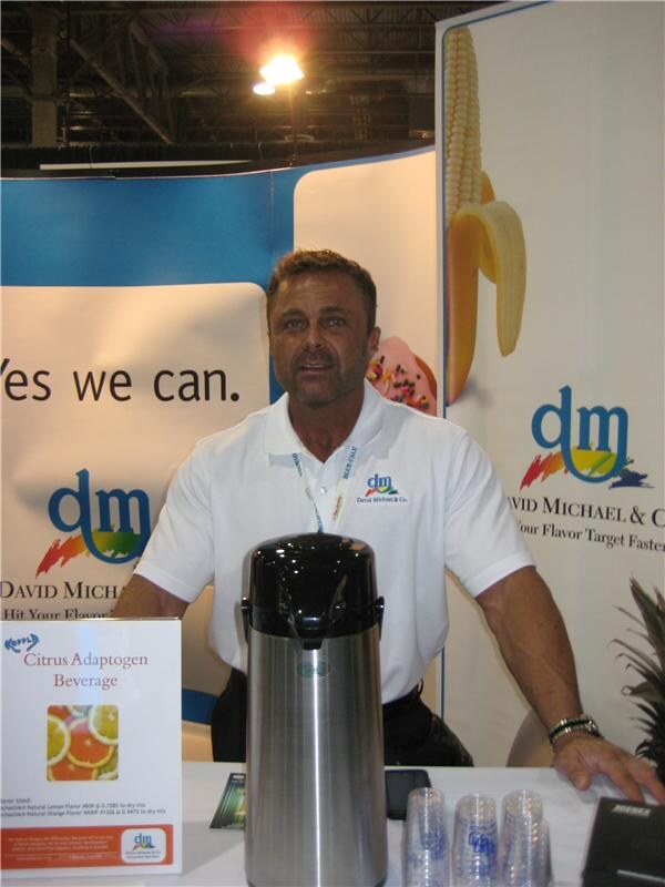 A man in a white David Michael & Co. polo stands behind a counter with a steel airpot and plastic cups, beside a sign for a citrus adaptogen beverage and banners reading 'Yes we can.'
