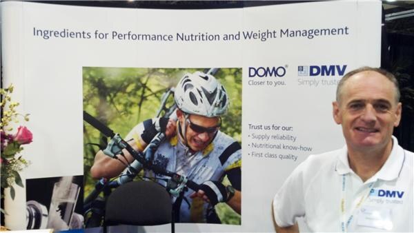A smiling man in a white DMV polo stands beside a DOMO and DMV banner showing a cyclist and the text 'Ingredients for Performance Nutrition and Weight Management'.