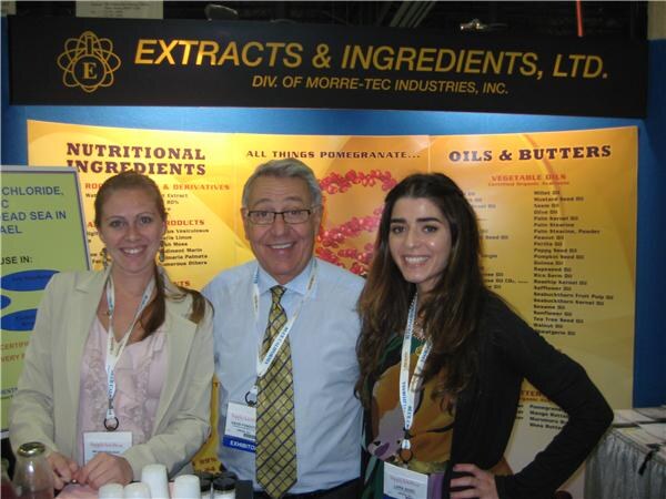 A man in glasses and a yellow tie stands between two smiling women wearing badges, in front of an Extracts & Ingredients, Ltd. sign and displays of nutritional ingredients and oils.