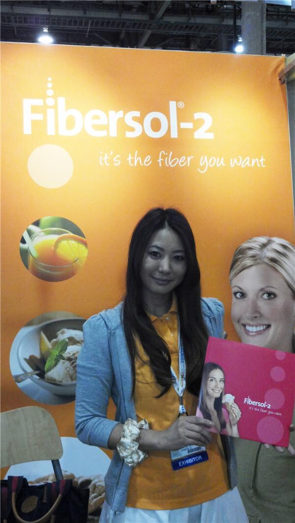 A woman with long dark hair and an exhibitor badge holds a pink Fibersol-2 brochure in front of an orange Fibersol-2 backdrop with food and drink images.