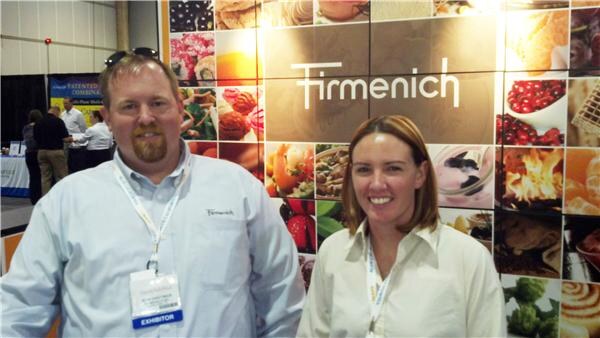 A bearded man with sunglasses on his head and a smiling woman stand in front of a Firmenich wall of food photographs, both wearing badges.
