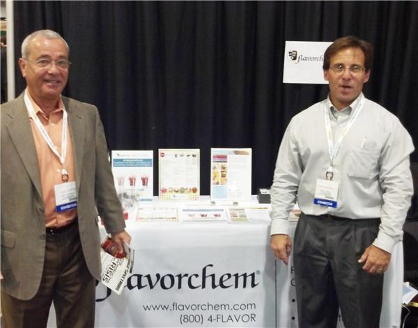 Two men wearing exhibitor badges stand on either side of a table with a Flavorchem tablecloth showing the website and phone number, with printed flyers on the table.