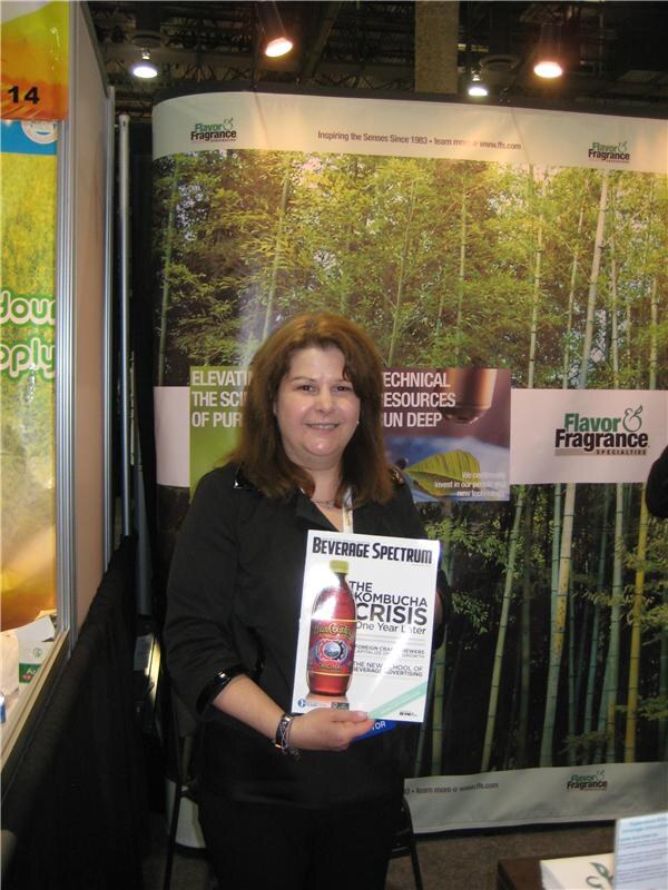 A woman with shoulder-length brown hair, dressed in black, holds a Beverage Spectrum magazine in front of a Flavor & Fragrance Specialties bamboo-forest banner.