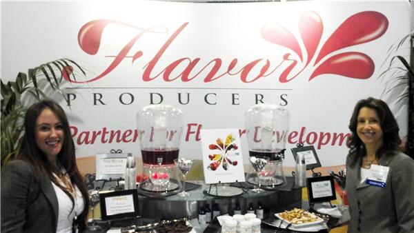 Two smiling women stand at either end of a display table in front of a large Flavor Producers banner, with two glass beverage dispensers, small bottles and snacks on the table.