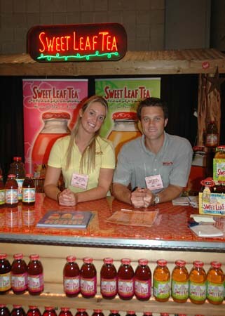 A woman and a man sit behind a counter under a lit Sweet Leaf Tea sign, with rows of bottled tea in front of them and banners behind.