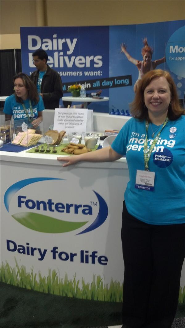 A smiling woman in a blue "I'm a morning person" T-shirt leans on a Fonterra counter. Behind her, another woman and a man stand at a display of breakfast foods under a "Dairy Delivers" banner.