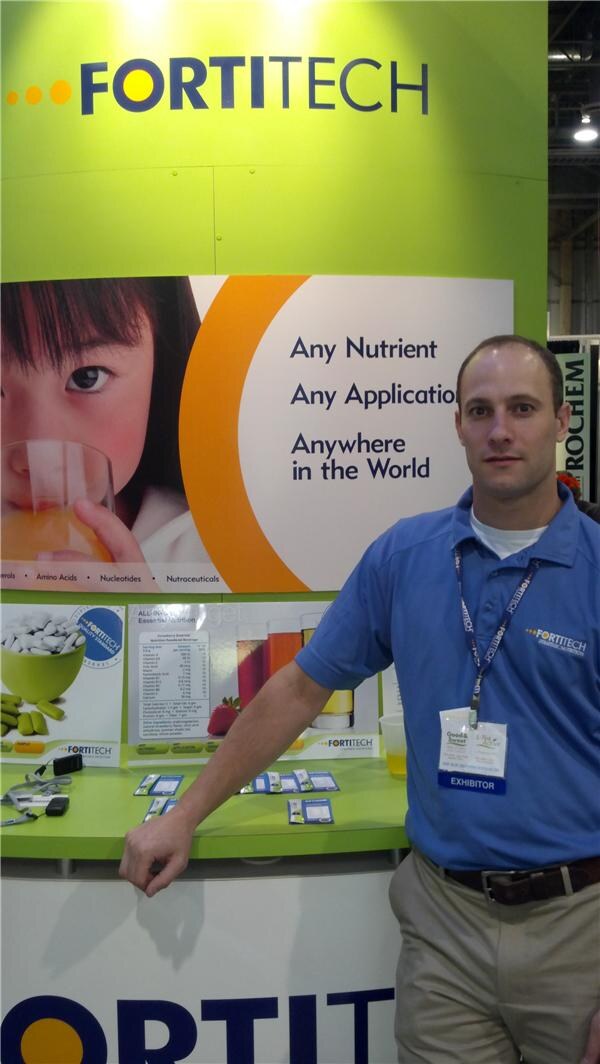 A man in a blue Fortitech polo and exhibitor badge leans on a green counter in front of a Fortitech display reading "Any Nutrient, Any Application, Anywhere in the World."