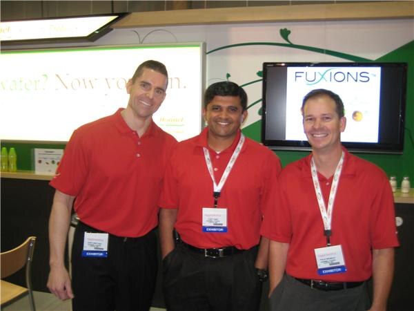 Three smiling men in red polo shirts, two wearing exhibitor badges, stand together in a booth with a screen showing the Fuxions logo behind them.