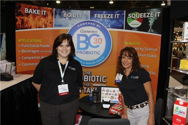 Two women in black Ganeden polo shirts stand in front of an orange Ganeden BC30 Probiotic banner reading "Bake it, Boil it, Freeze it, Squeeze it."