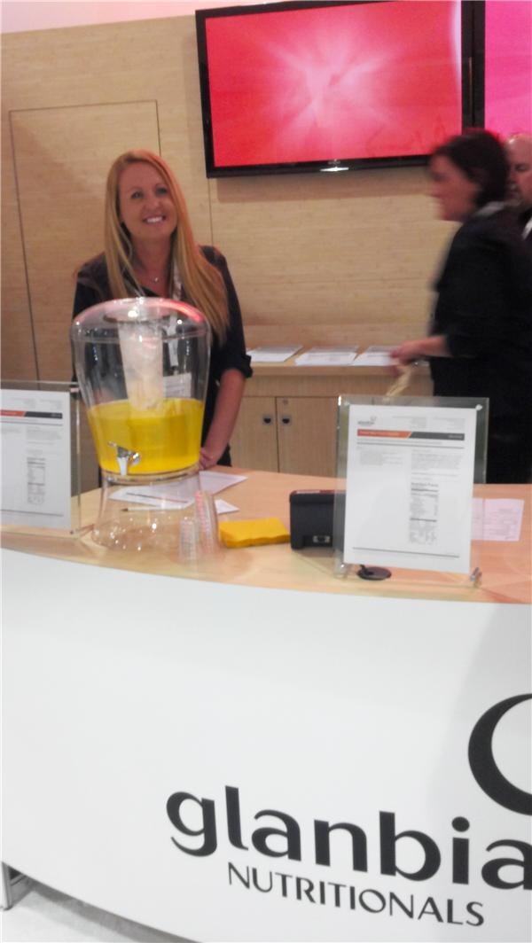 A smiling woman with long blonde hair stands behind a Glanbia Nutritionals counter holding a glass dispenser of yellow drink, with printed sheets on stands and a red screen behind her.