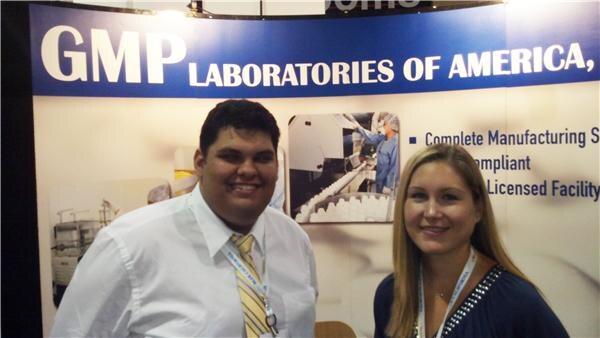 A smiling man in a white shirt and yellow striped tie stands beside a woman with long blonde hair in front of a GMP Laboratories of America banner.