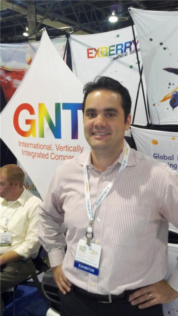 A smiling man with dark hair, in a checked shirt with an exhibitor badge and lanyard, stands with a hand on his hip in front of GNT and Exberry banners. A seated man is partly visible at left.