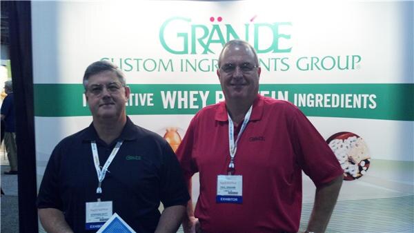 Two men with exhibitor badges stand side by side, one in a black polo and one in a red polo, in front of a Grande Custom Ingredients Group banner about whey protein ingredients.