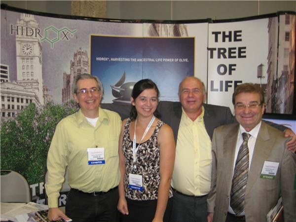 Three men and a woman stand close together at a Hidrox booth with a banner reading The Tree of Life and a city skyline backdrop.