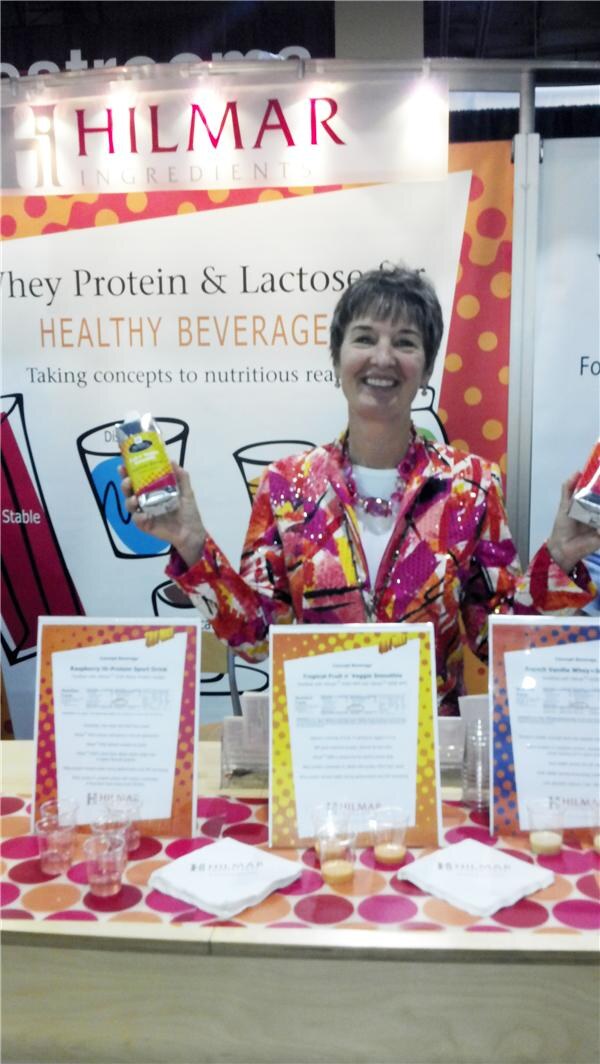 A smiling woman in a colorful patterned jacket holds up a beverage package in each hand behind a table of sample cups and concept beverage signs under a Hilmar Ingredients banner.