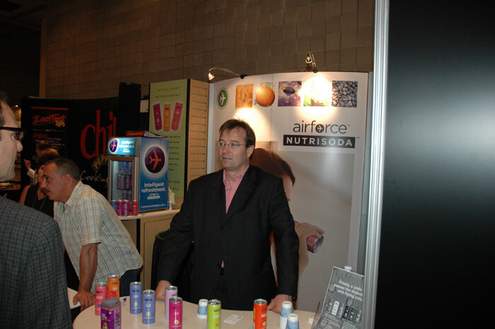 A man in a dark jacket stands behind a round table of colorful Airforce Nutrisoda cans, beside a small cooler and a banner. Other attendees stand nearby.