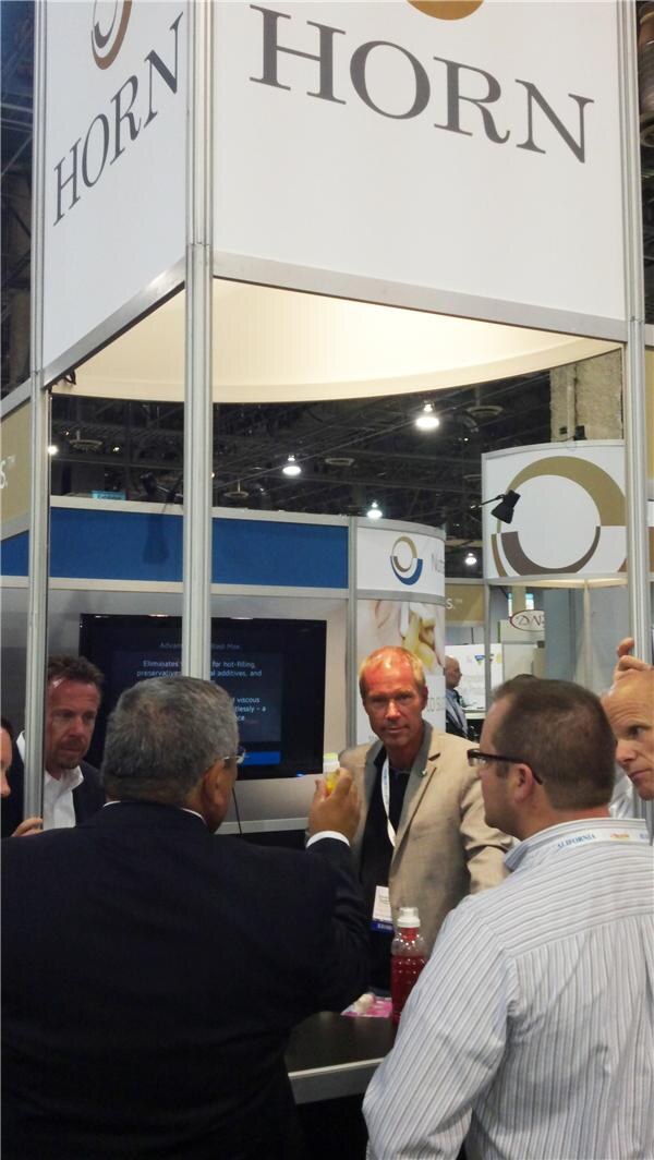 Several men in business attire talk at a booth under large Horn signs. One holds a small bottle, and a bottle of red liquid sits on the counter in front of a screen.