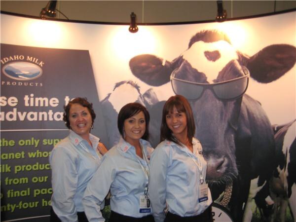 Three smiling women in light blue shirts with badges stand in front of an Idaho Milk Products display featuring a large image of a cow wearing sunglasses.