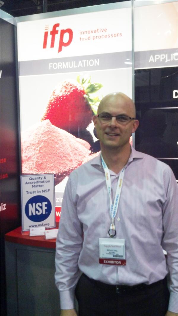 A bald man with glasses and an exhibitor badge stands in front of an ifp innovative food processors display showing a strawberry and powder, next to an NSF sign.
