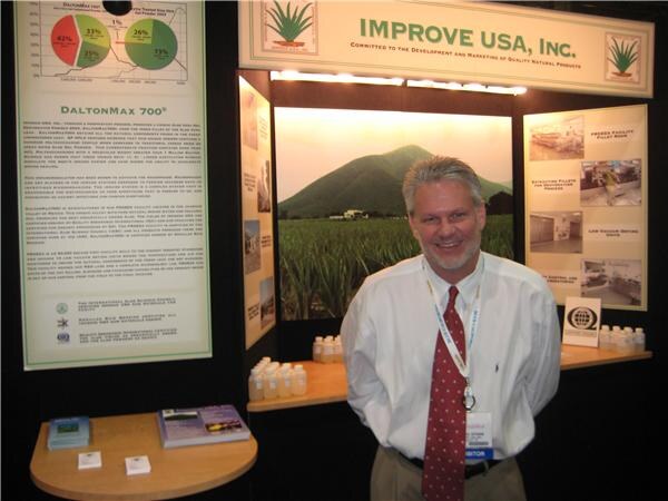 A smiling gray-haired man in a white shirt and red tie stands in the Improve USA, Inc. booth, with a DaltonMax 700 poster, a field photo and product bottles behind him.