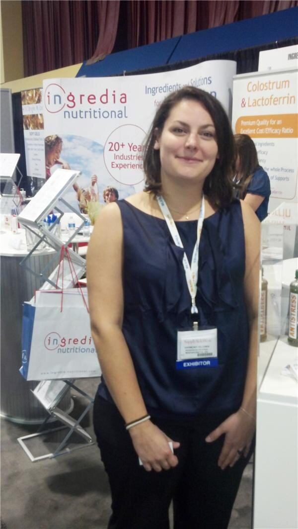 A woman with dark hair in a navy sleeveless top and exhibitor badge stands in front of Ingredia Nutritional banners and a literature stand with a shopping bag.