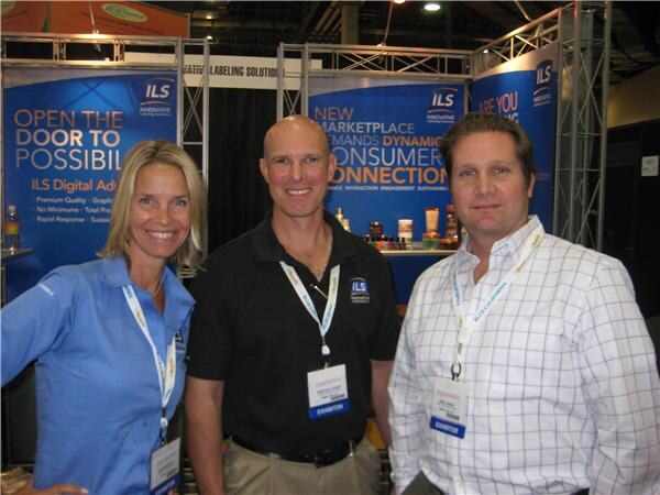 Two men and a woman, all wearing exhibitor badges, pose together in front of blue Innovative Labeling Solutions (ILS) banners at the booth.
