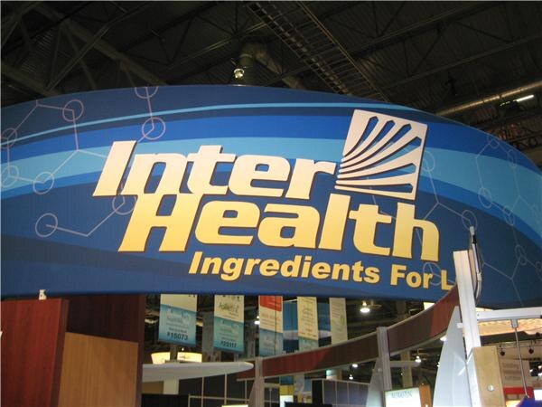 A large curved blue overhead sign reading InterHealth Ingredients For L, with a sunburst logo, hangs above the InterHealth exhibit on the show floor.