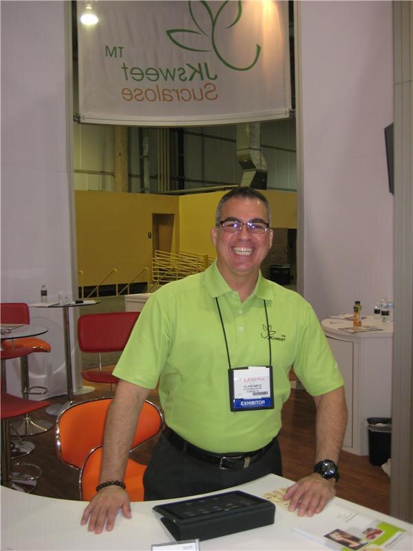 A smiling man with glasses in a green JKsweet polo and exhibitor badge stands behind a white counter at the JKsweet Sucralose booth, with orange and red chairs behind him.