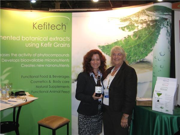 Two women stand together in front of a green Kefitech display; the woman on the left holds a bottle. Brochures sit on a table with a green cloth.