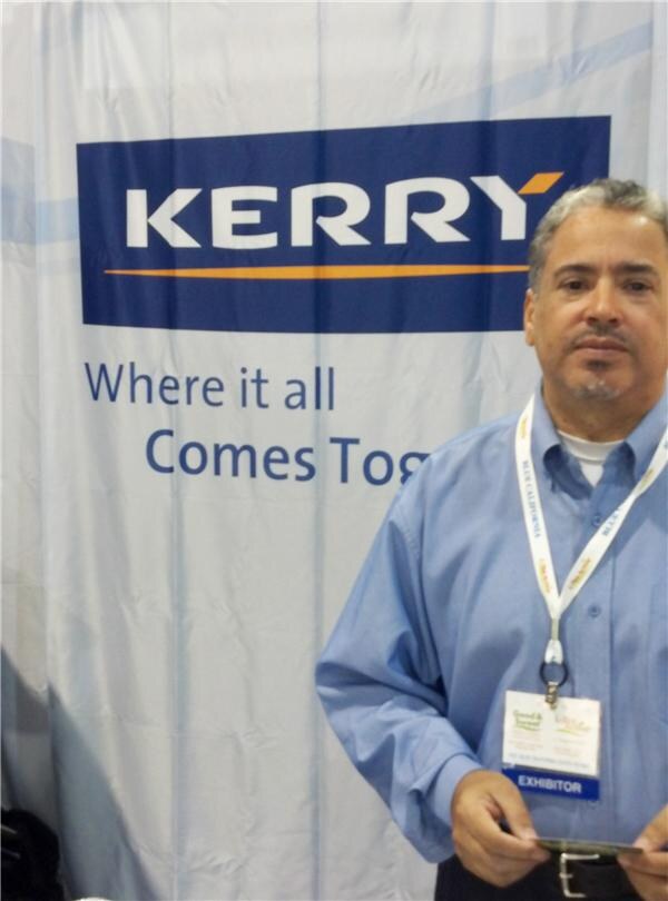 A man in a light blue shirt with a lanyard and exhibitor badge stands in front of a Kerry banner reading Where it all Comes Together.