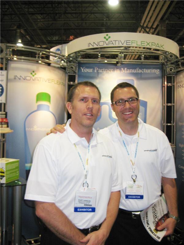 Kurt Hanson of Innovative Flexpak, wearing a white polo and exhibitor badge, stands beside a smiling man with glasses in front of the Innovative Flexpak booth.