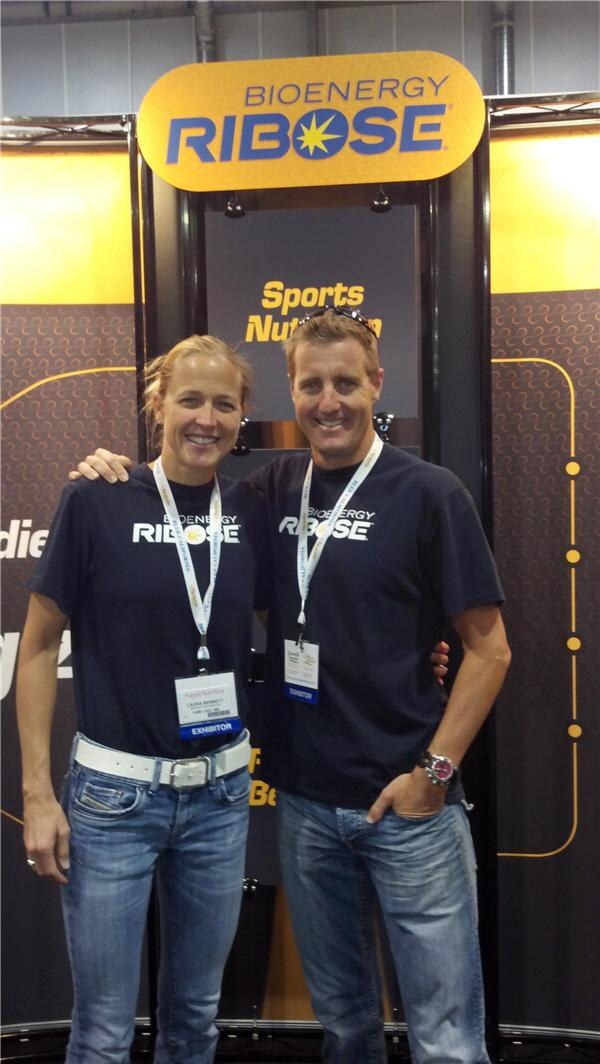 Laura Bennett, at left, and a man in matching black Bioenergy Ribose shirts smile with arms around each other in front of the Bioenergy Ribose sign. Both wear exhibitor badges.