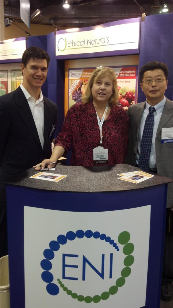 Lauren Clancy stands at center behind a counter with the ENI logo, flanked by a man in a dark suit and a man in glasses and a tie, under an Ethical Naturals sign.