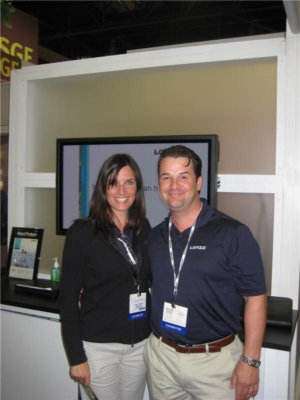 A smiling woman in a black jacket and a man in a navy Lonza polo, both wearing exhibitor badges, stand together in front of a monitor at the Lonza booth.
