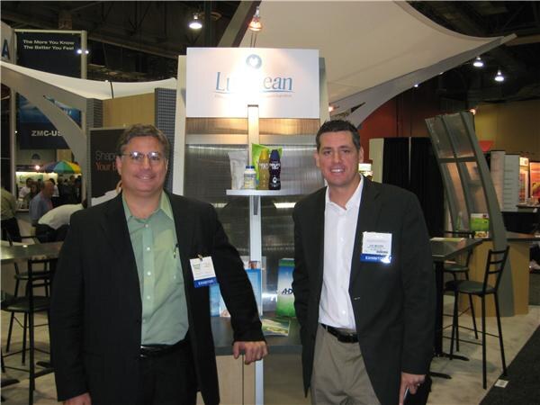 Two men in dark jackets with exhibitor badges stand beside a Lumilean display holding packaged products, in a trade-show hall.