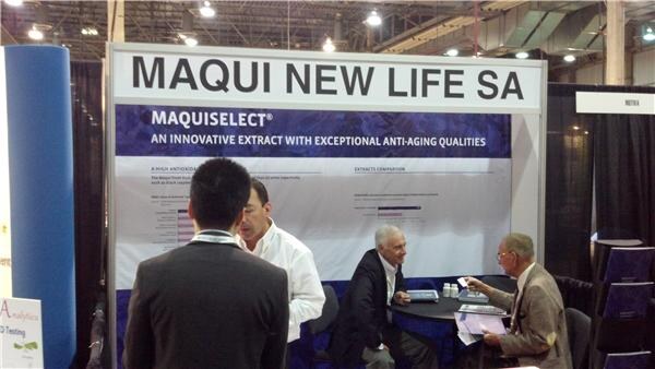 Booth with a Maqui New Life SA sign and a MaquiSelect display; two seated men talk at a table while a man in a suit jacket stands with his back to the camera.