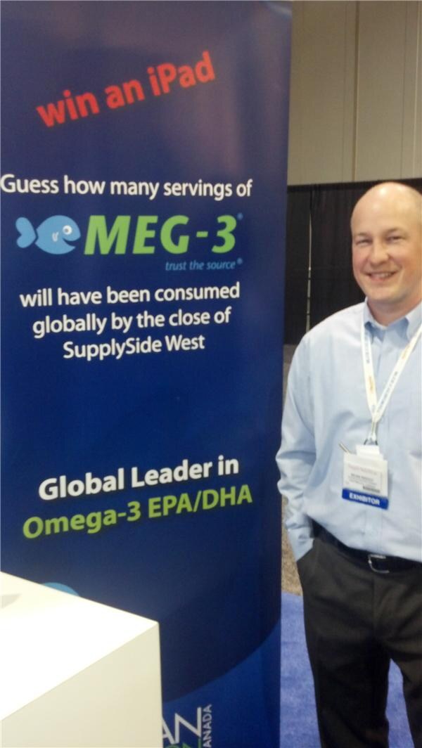 A smiling bald man in a light blue shirt with an exhibitor badge stands beside a blue MEG-3 banner offering a chance to win an iPad and describing omega-3 EPA/DHA.