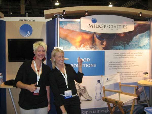 Two women in black tops and exhibitor badges smile at the Milk Specialties Global booth; one points toward a Food Solutions panel showing a swimmer.